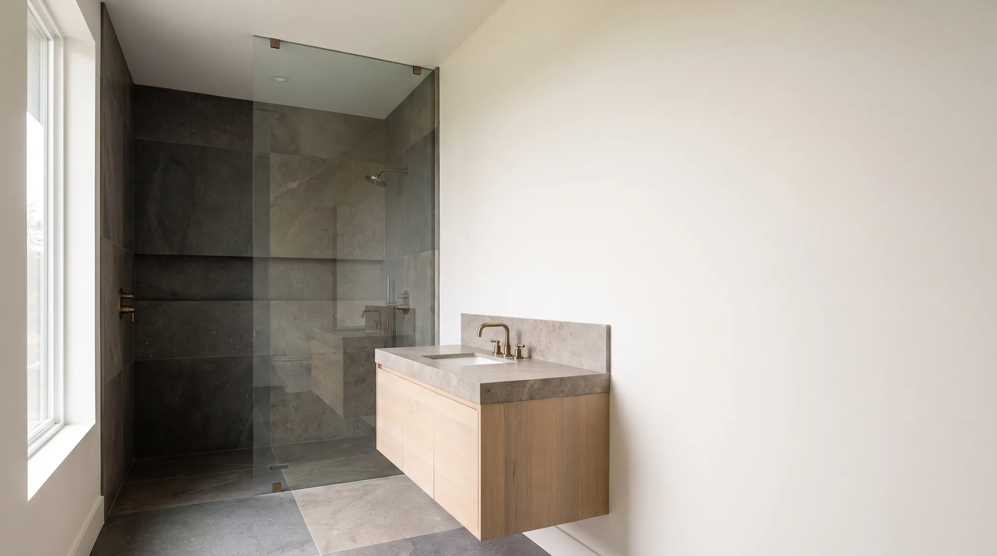 A finished bathroom with a tiled walk-in shower, floating oak vanity and warm brass fixtures in soft daylight